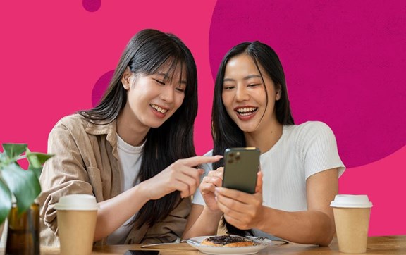Coppia di amiche asiatiche al tavolo di un fastfood che guardano insieme uno smartphone ridendo.
