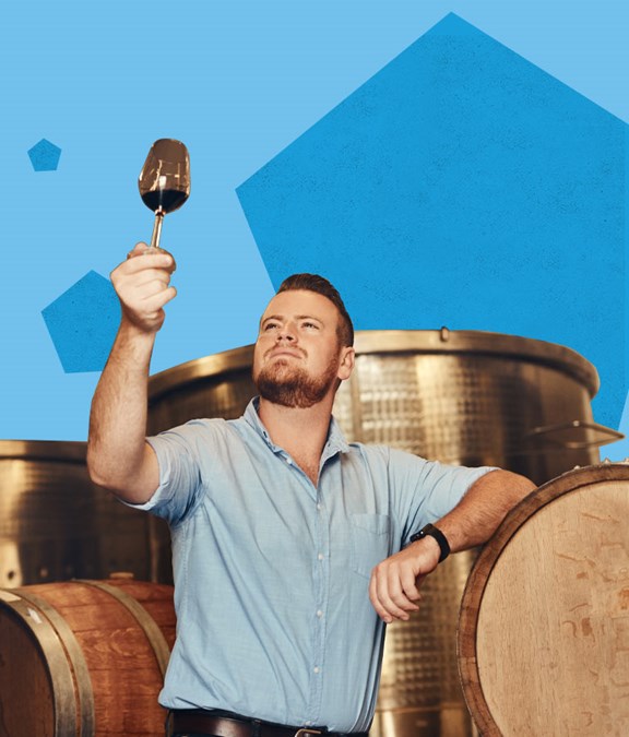 Ragazzo che guarda concentrato il colore del vino all'interno del calice tenuto in alto dalla mano... appoggiato a delle botti all'interno di una cantina.