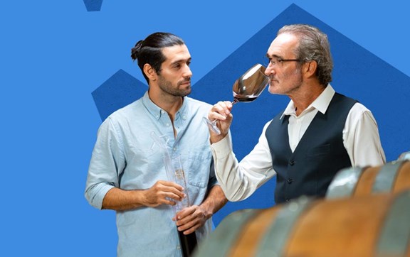 Un ragazzo con in mano una ampolla molto grande guarda un signore che odora un calice di vino rosso all'interno di una cantina, ci sono le botti.