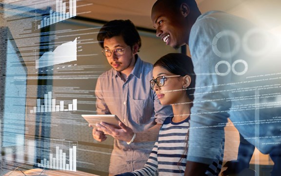 Tre persone, una ragazza seduta davanti a un PC e due ragazzi in piedi al suo fianco che guardano tutti insieme un monitor.